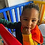 toddler, child, curly_hair, teething, playpen, colorful, red, blue, yellow, fingers_in_mouth, indoor, toy, window, natural_light, clothing, orange, yellow_sleeve, green_sleeve, face, person