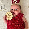 baby, infant, child, red_dress, rose_pattern, milestone_card, crown, smiling, lying_down, blanket, happy, cute, portrait, hand, face, blue_eyes, skin, celebration, age_marker, indoors