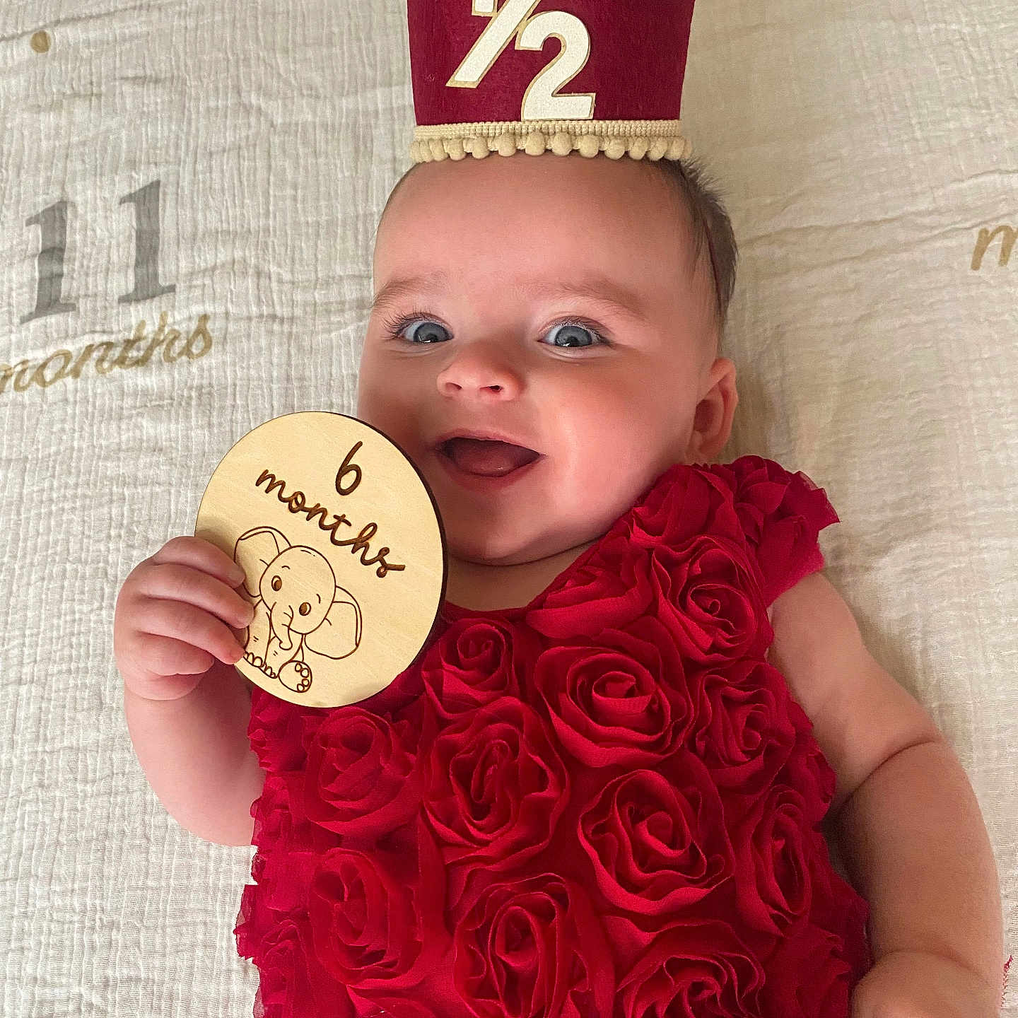 Adeline joined the competition — help win amazing prizes! baby, infant, child, red_dress, rose_pattern, milestone_card, crown, smiling, lying_down, blanket, happy, cute, portrait, hand, face, blue_eyes, skin, celebration, age_marker, indoors