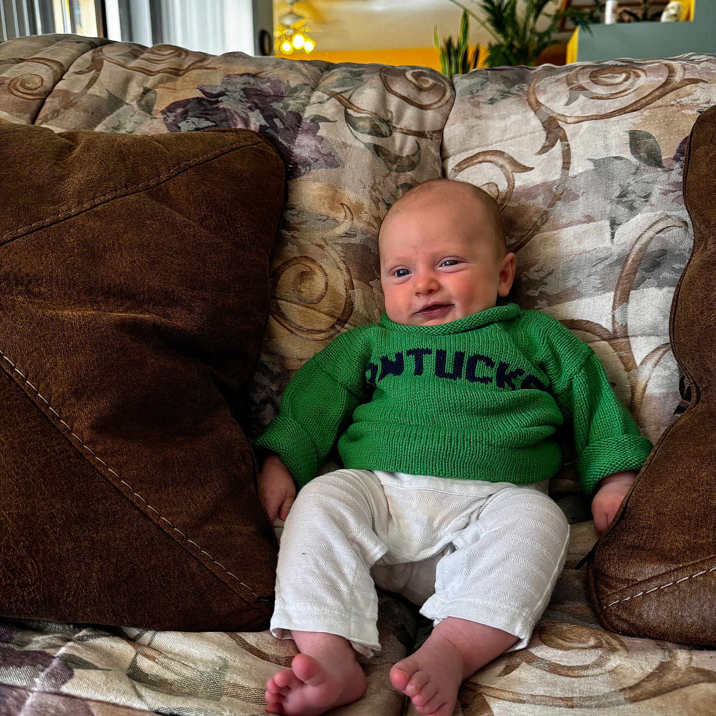 Eaton joined the competition — help win amazing prizes! baby, child, couch, cozy, cute, face, feet, furniture, green_sweater, home, indoor, infant, living_room, person, pillows, portrait, relaxed, sitting, smiling, white_pants