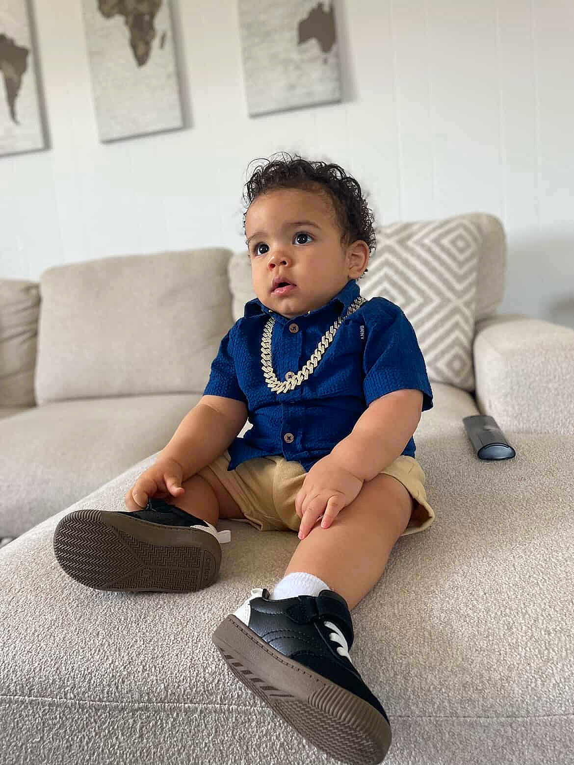 Kenny joined the competition — help win amazing prizes! toddler, child, sitting, couch, blue_shirt, shorts, sneakers, gold_chain, curly_hair, indoor, living_room, cushion, wall_art, map, footwear, person, cute, portrait, casual_clothing, neutral_colors