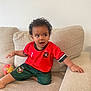 Kenny joined the competition — help win amazing prizes! beige_couch, casual, child, colorful_toy, couch, curious, curly_hair, green_shorts, happy, home, indoor, person, portrait, red_shirt, relaxed, sitting, soccer_uniform, toddler, toy, young_child