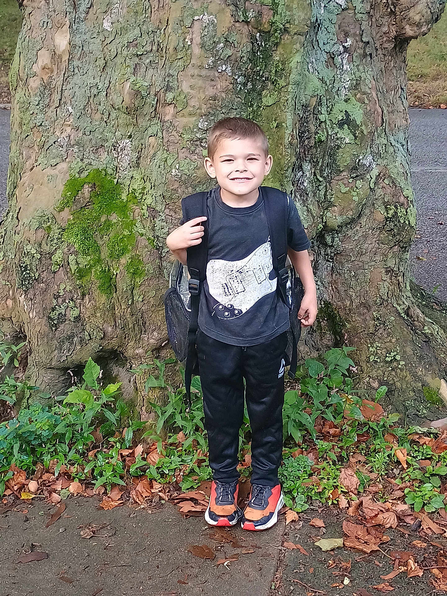 Nickalus is registered to the contest to win money with this photo: child, boy, tree, backpack, tshirt, guitar_print, pants, sneakers, smile, outdoor, nature, leaves, sidewalk, greenery, casual_clothing, happy, standing, daytime, park, young
