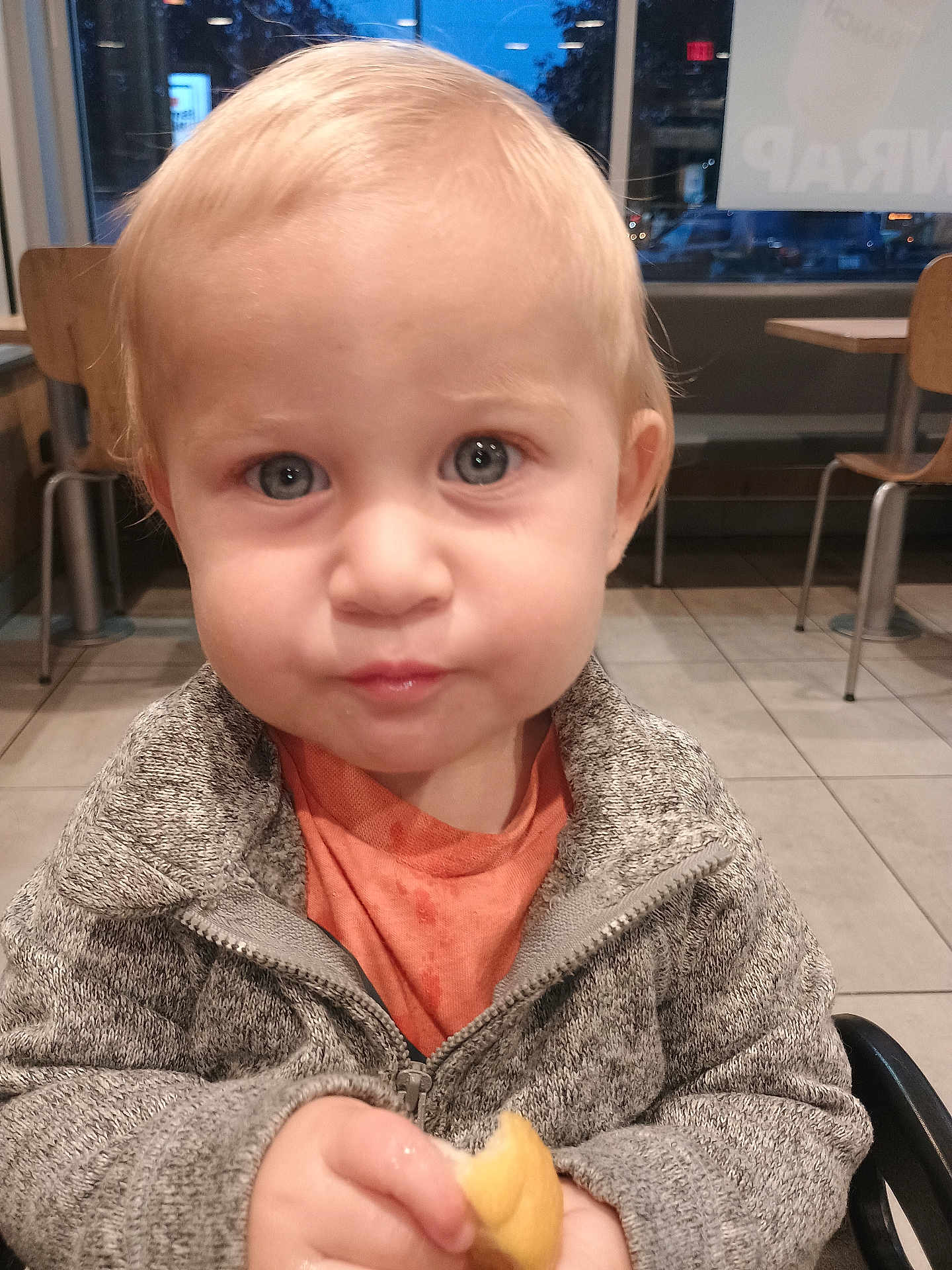 Tristen is registered to the contest to win money with this photo: toddler, child, indoor, face, blue_eyes, clothing, gray_jacket, orange_shirt, bread, food, sitting, floor, wooden_chair, table, window, curious, portrait, young_child, cute, casual
