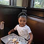 child, smiling, curly_hair, white_shirt, messy_shirt, restaurant, booth, tray, food_tray, sauce_cup, plastic_cup, table, window, daylight, indoor, person, seat, happy, curly_ponytail, eating