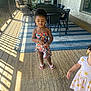 Eliyah is registered to the contest to win money with this photo: child, girl, floral_dress, porch, rug, sunlight, shadow, patio_furniture, table, chair, outdoor, wood_floor, barefoot, curly_hair, colorful_clothing, summer, daylight, holding_object, young_child, casual
