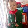 baby, child, elf_outfit, green_clothing, red_clothing, smiling, indoor, table, storage_boxes, picture_frame, curtains, floral_pattern, person, furniture, portrait, holiday_costume, cute, seated, home, natural_light