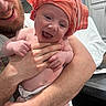 baby, infant, smile, happy, adult, man, beard, hand, holding, diaper, orange_cloth, headwear, indoor, kitchen, cabinet, countertop, skin, person, closeup, affection