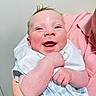 Hank is registered to the contest to win money with this photo: baby, smiling, blue_eyes, infant, happy, person, holding, arm, clothing, bib, pastel_colors, pink_shirt, checkered_pattern, skin, indoor, portrait, cute, newborn, face, closeup