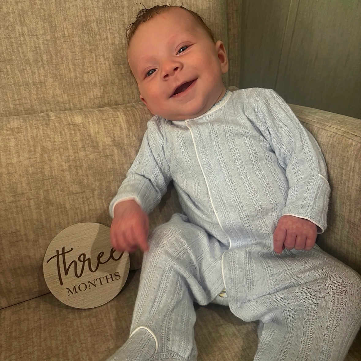 Hank is registered to the contest to win money with this photo: baby, smile, infant, blue_onesie, couch, knitted_clothing, milestone_sign, wooden_sign, sitting, happy, child, indoor, portrait, face, person, cute, young_child, three_months, cozy, furniture