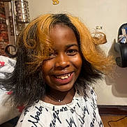 Mahraj joined the competition — help win amazing prizes! child, smile, hair, two_tone_hair, indoor, shirt, text_on_clothing, necklace, face, happy, lamp, chair, wooden_floor, person, portrait, casual, home, cozy, black_hair, blonde_hair