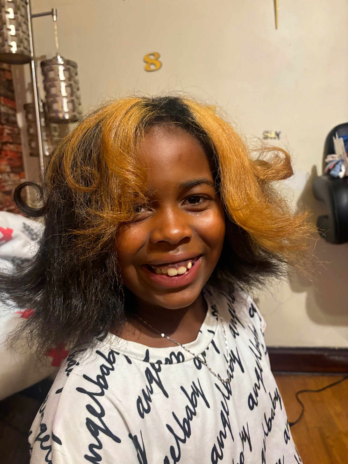 Mahraj joined the competition — help win amazing prizes! child, smile, hair, two_tone_hair, indoor, shirt, text_on_clothing, necklace, face, happy, lamp, chair, wooden_floor, person, portrait, casual, home, cozy, black_hair, blonde_hair