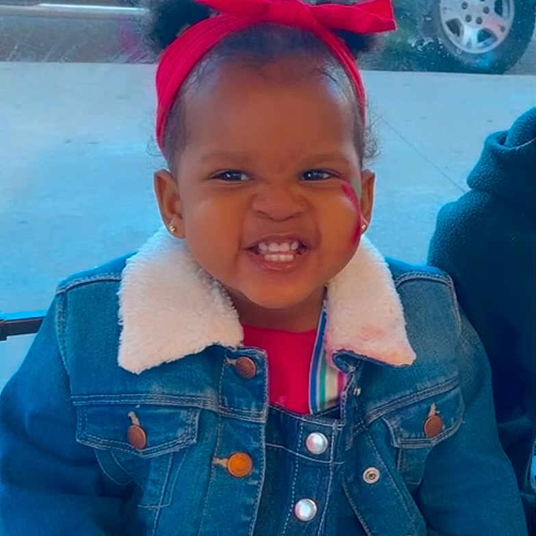 Amorah joined the competition — help win amazing prizes! button, child, clothing, cute, denim_jacket, expression, face_paint, happy, indoor, jacket, person, playful, portrait, red_headband, sitting, smile, toddler, vehicle_wheel, window, young_child