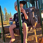 Tony is registered to the contest to win money with this photo: toddler, child, playground, swing, snack, sippy_cup, cap, backpack, trees, sunlight, outdoor, woodchips, play_equipment, fence, summer, casual_clothing, barefoot, daytime, happy, park