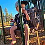 toddler, child, playground, swing, snack, sippy_cup, cap, backpack, trees, sunlight, outdoor, woodchips, play_equipment, fence, summer, casual_clothing, barefoot, daytime, happy, park