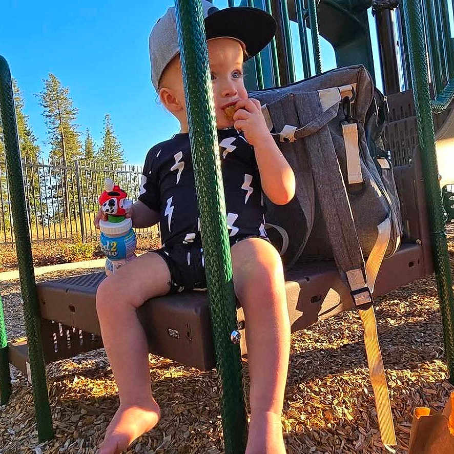Tony is registered to the contest to win money with this photo: backpack, barefoot, cap, casual_clothing, child, daytime, fence, happy, outdoor, park, play_equipment, playground, sippy_cup, snack, summer, sunlight, swing, toddler, trees, woodchips