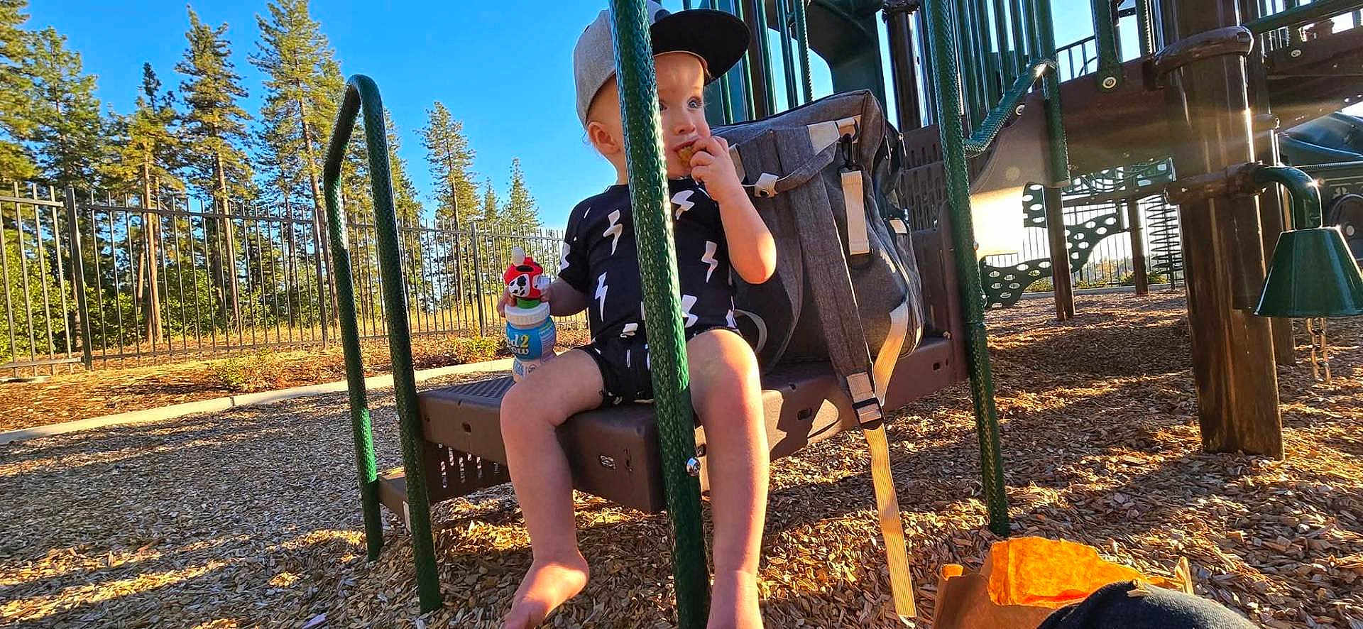 Tony is registered to the contest to win money with this photo: toddler, child, playground, swing, snack, sippy_cup, cap, backpack, trees, sunlight, outdoor, woodchips, play_equipment, fence, summer, casual_clothing, barefoot, daytime, happy, park