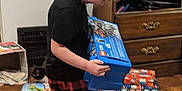 James is registered to the contest to win money with this photo: child, pajamas, lego, toy_box, wrapping_paper, floor, cat, box, dresser, television, room, wooden_floor, unwrapped_presents, plaid, black_shirt, excitement, holiday, gift, indoor, person