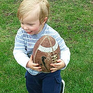 James is registered to the contest to win money with this photo: toddler, child, football, grass, outdoor, sport, happy, smiling, playing, clothing, striped_shirt, blue_pants, toy, active, cute, young, boy, fun, daylight, casual