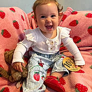 Delilah joined the competition — help win amazing prizes! baby, child, smiling, happy, indoor, plush_toy, pink_blanket, strawberry_pattern, clothing, pants, shoes, milestone_card, person, cute, young_child, sitting, face, hair, room, toy