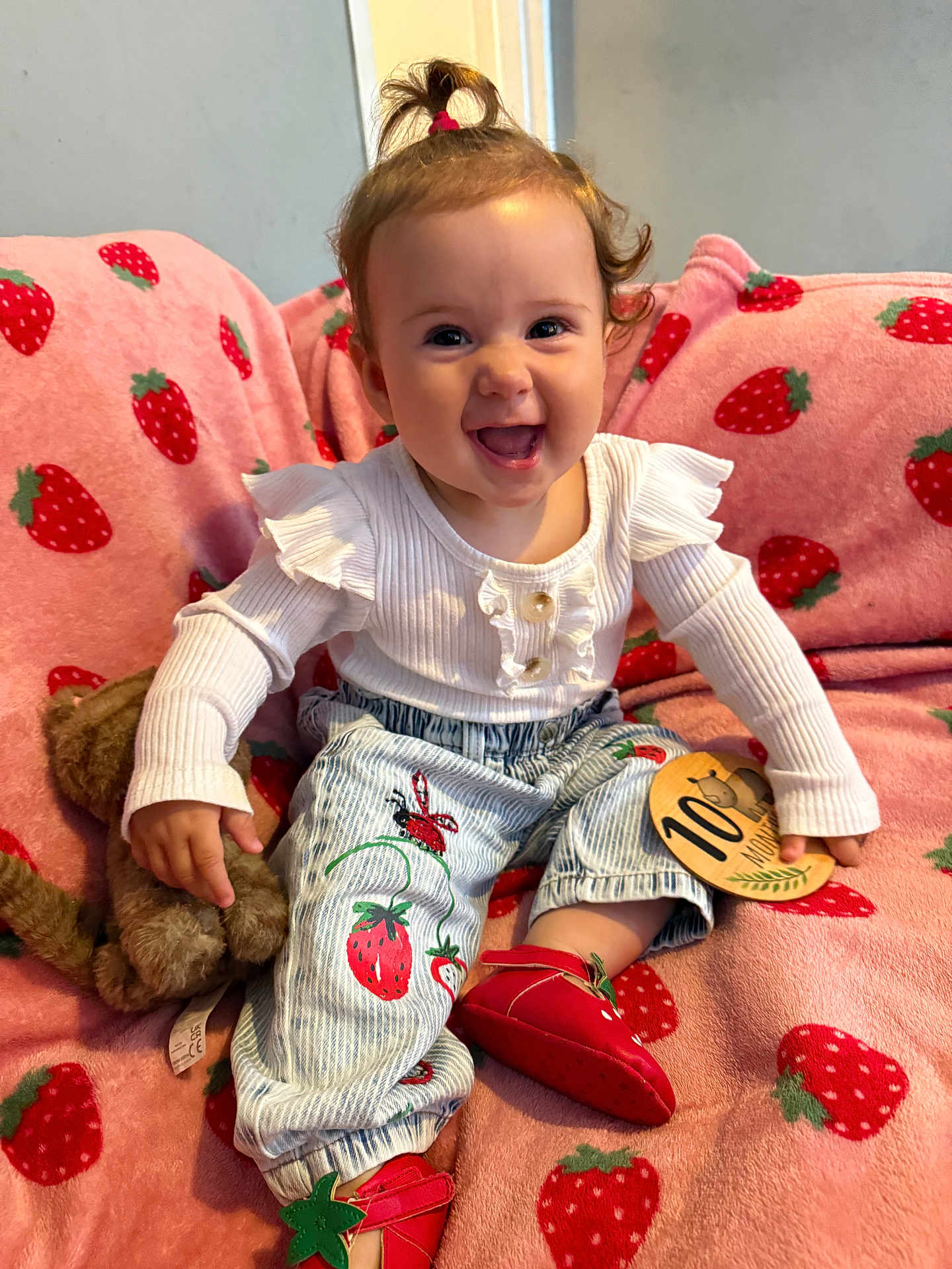 Delilah joined the competition — help win amazing prizes! baby, child, smiling, happy, indoor, plush_toy, pink_blanket, strawberry_pattern, clothing, pants, shoes, milestone_card, person, cute, young_child, sitting, face, hair, room, toy