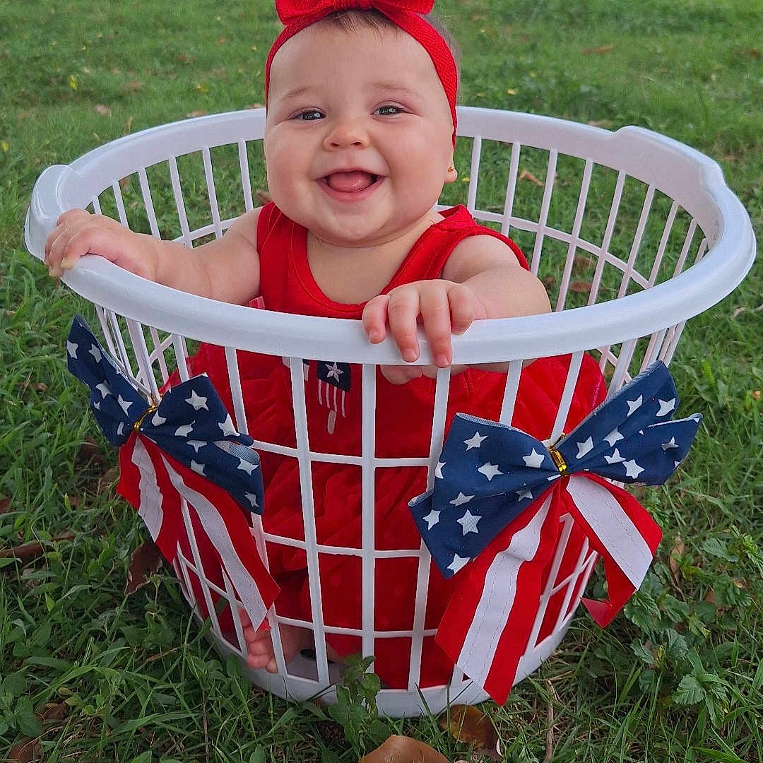 Allysson is registered to the contest to win money with this photo: american_flag, baby, bow, celebration, child, cute, grass, happy, headband, holiday, infant, laundry_basket, nature, outdoor, patriotic, person, playful, red_dress, sitting, smiling