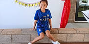 Maryfer is registered to the contest to win money with this photo: banner, bench, blue_jersey, brick_wall, child, concrete_floor, crocs, decoration, indoor, playful, portrait, red_curtain, school_event, sitting, smile, sports_uniform, welcome_sign, white_shoes, window, wooden_bench