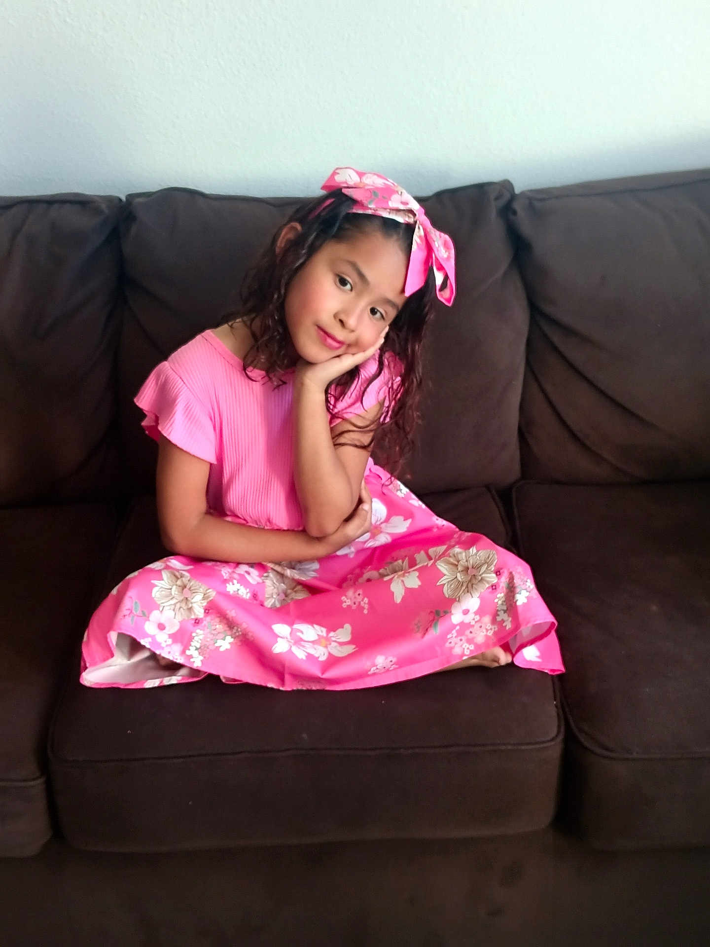 Maryfer is registered to the contest to win money with this photo: child, girl, pink_dress, headband, couch, sitting, portrait, smile, long_hair, indoor, relaxed, brown_couch, floral_pattern, casual, cute, young, resting_hand, fashion, home, happy