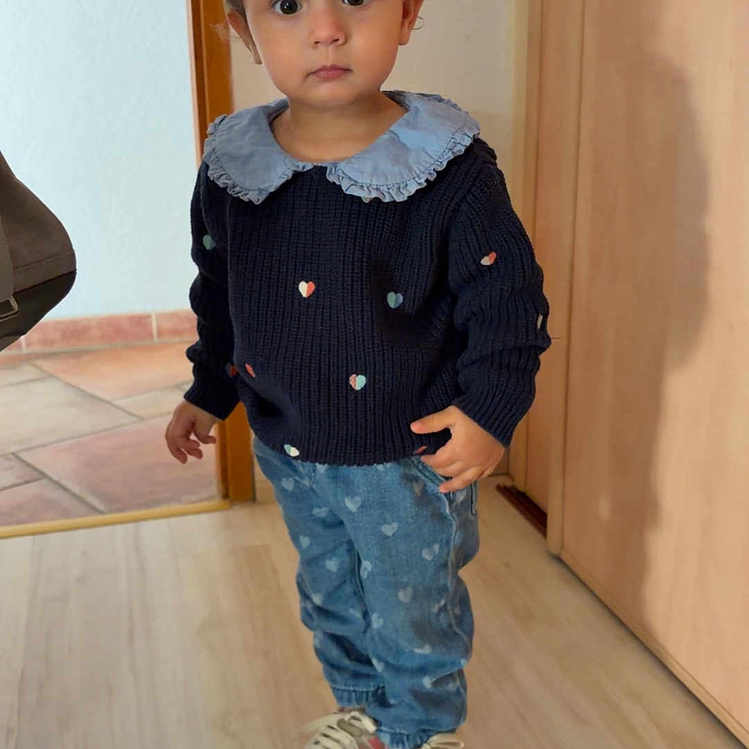 Giorgia participe au concours pour gagner de l'argent avec cette photo : casual_clothing, child, curious, door, fashion, floor, full_body, heart_pattern, indoor, jeans, light_wood_floor, neutral_expression, person, portrait, sneakers, standing, sweater, toddler, wall, young_child