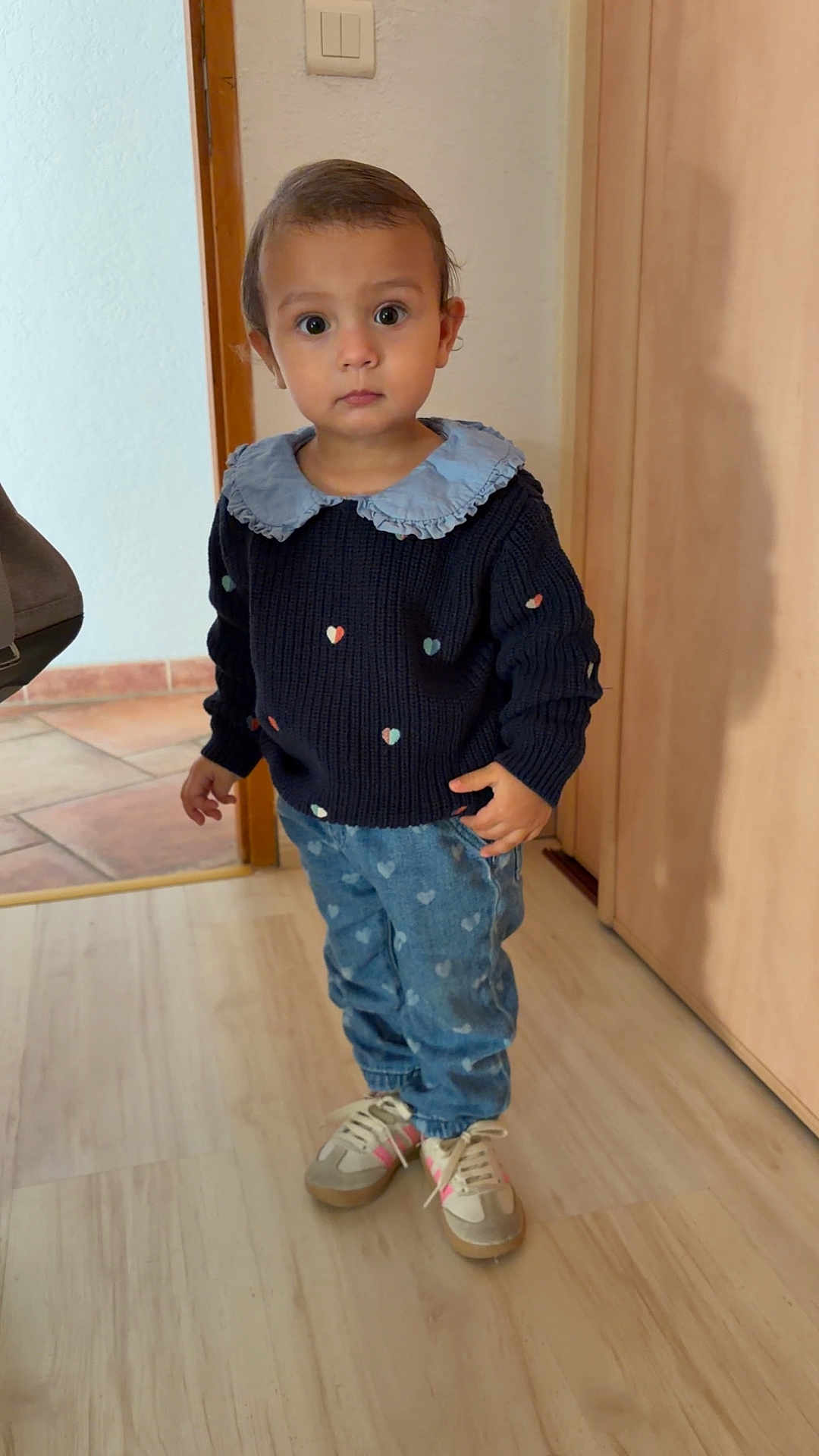 Giorgia participe au concours pour gagner de l'argent avec cette photo : toddler, child, indoor, floor, sweater, jeans, sneakers, heart_pattern, curious, standing, person, young_child, casual_clothing, light_wood_floor, door, wall, neutral_expression, portrait, full_body, fashion