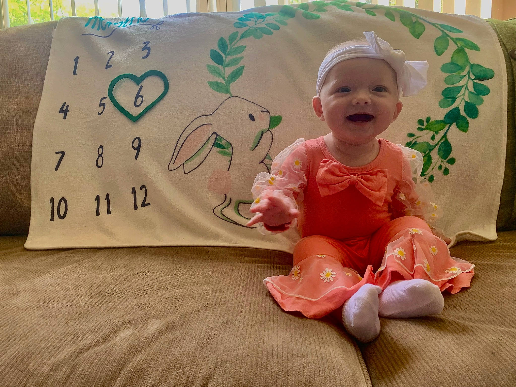 Bailey is registered to the contest to win money with this photo: baby, baby_toddler_clothing, bedding, child, comfort, fashion_accessory, flooring, font, foot, happy, linens, peach, person, room, sitting, sleeve, smile, textile, throw_pillow, toddler