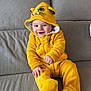 baby, child, yellow_onesie, hood, smiling, couch, indoor, plush_clothing, happy, cute, infant, seated, face, portrait, cozy, home, furniture, hands, feet, animal_costume