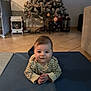 baby, child, floor, mat, christmas_tree, holiday, indoor, smiling, face, person, decorations, festive, home, couch, tiled_floor, toy, lamp, cozy, striped_shirt, hands