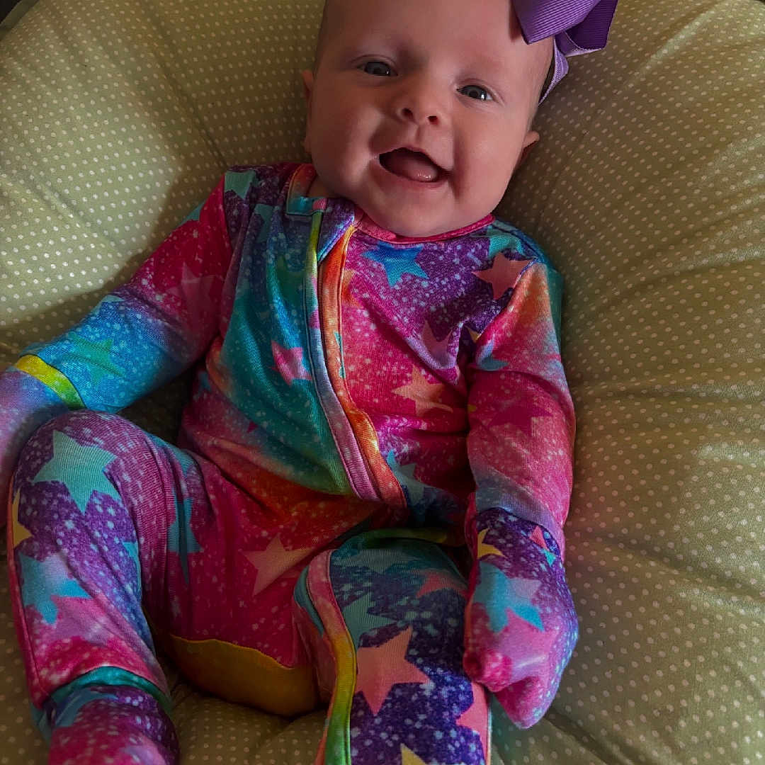 Ella joined the competition — help win amazing prizes! baby, baby_clothes, child, colorful_clothing, cozy, cushion, cute, face, happy, headwear, indoors, infant, onesie, polka_dots, portrait, purple_bow, sitting, smile, soft_lighting, stars_pattern