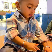 Isaac is registered to the contest to win money with this photo: toddler, child, plaid_shirt, black_pants, sitting, table, indoor, curious, bangle, shoe, modern_interior, blue_chair, light_decoration, person, portrait, casual_clothing, bottle, hand, face, young_child