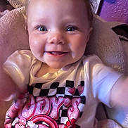 Drew is registered to the contest to win money with this photo: baby, child, smile, face, infant, clothing, tshirt, blanket, indoors, portrait, cute, happy, person, pink, purple, cheerful, young, expression, sitting, cozy