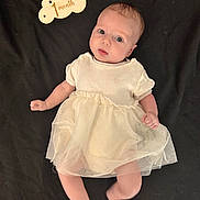 Lyla participe au concours pour gagner de l'argent avec cette photo : baby, infant, child, dress, cream_dress, lying_down, black_background, one_month, sign, cloud_sign, cute, young_child, portrait, indoors, person, newborn, face, eyes, tongue_out, soft_lighting