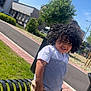 bench, blue_sky, boy, building, casual_clothing, child, curly_hair, daytime, grass, happy, outdoor, person, shorts, sidewalk, smiling, street, sunny, tree, tshirt, urban