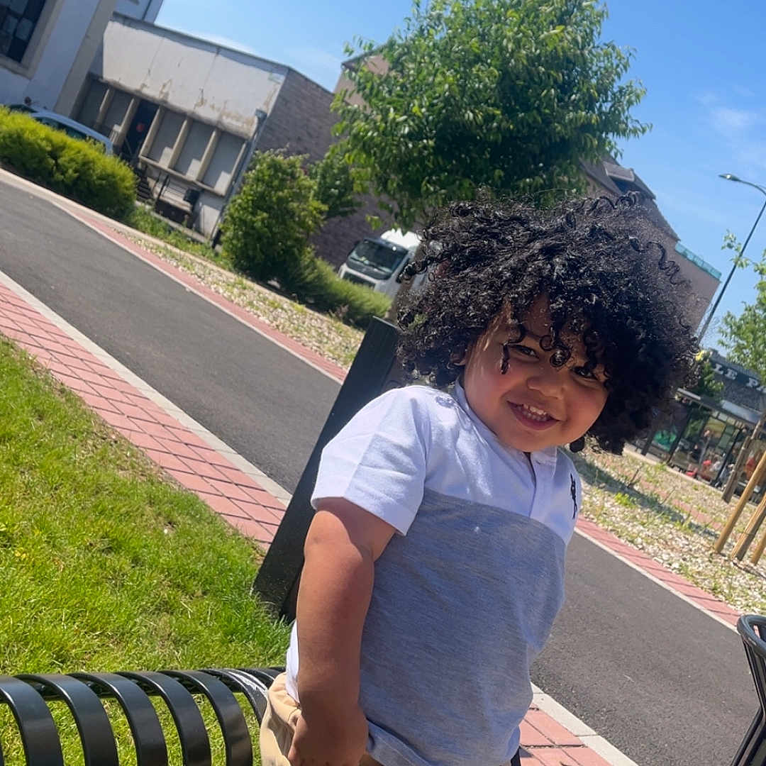 Junayd a rejoint le concours — aidez-le/la à gagner de superbes lots ! bench, blue_sky, boy, building, casual_clothing, child, curly_hair, daytime, grass, happy, outdoor, person, shorts, sidewalk, smiling, street, sunny, tree, tshirt, urban