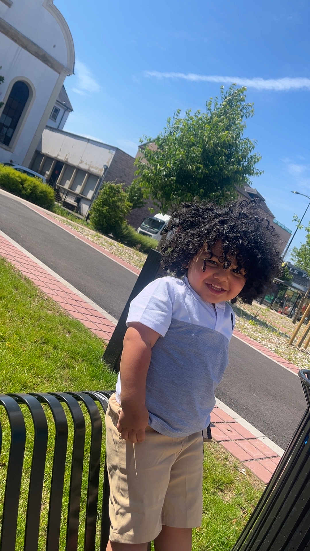 Junayd a rejoint le concours — aidez-le/la à gagner de superbes lots ! bench, blue_sky, boy, building, casual_clothing, child, curly_hair, daytime, grass, happy, outdoor, person, shorts, sidewalk, smiling, street, sunny, tree, tshirt, urban