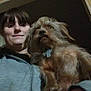 person, dog, indoor, selfie, hoodie, brown_dog, wooden_wall, doorframe, pet, fur, hand, casual_clothing, animal, portrait, face, canine, companion, holding, dim_lighting, neutral_expression