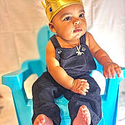Kylian is registered to the contest to win money with this photo: baby, child, crown, blue_chair, overalls, jewelry, golden_crown, portrait, cute, sitting, person, indoor, infant, baby_clothes, bracelet, necklace, expression, skin, foot, hand
