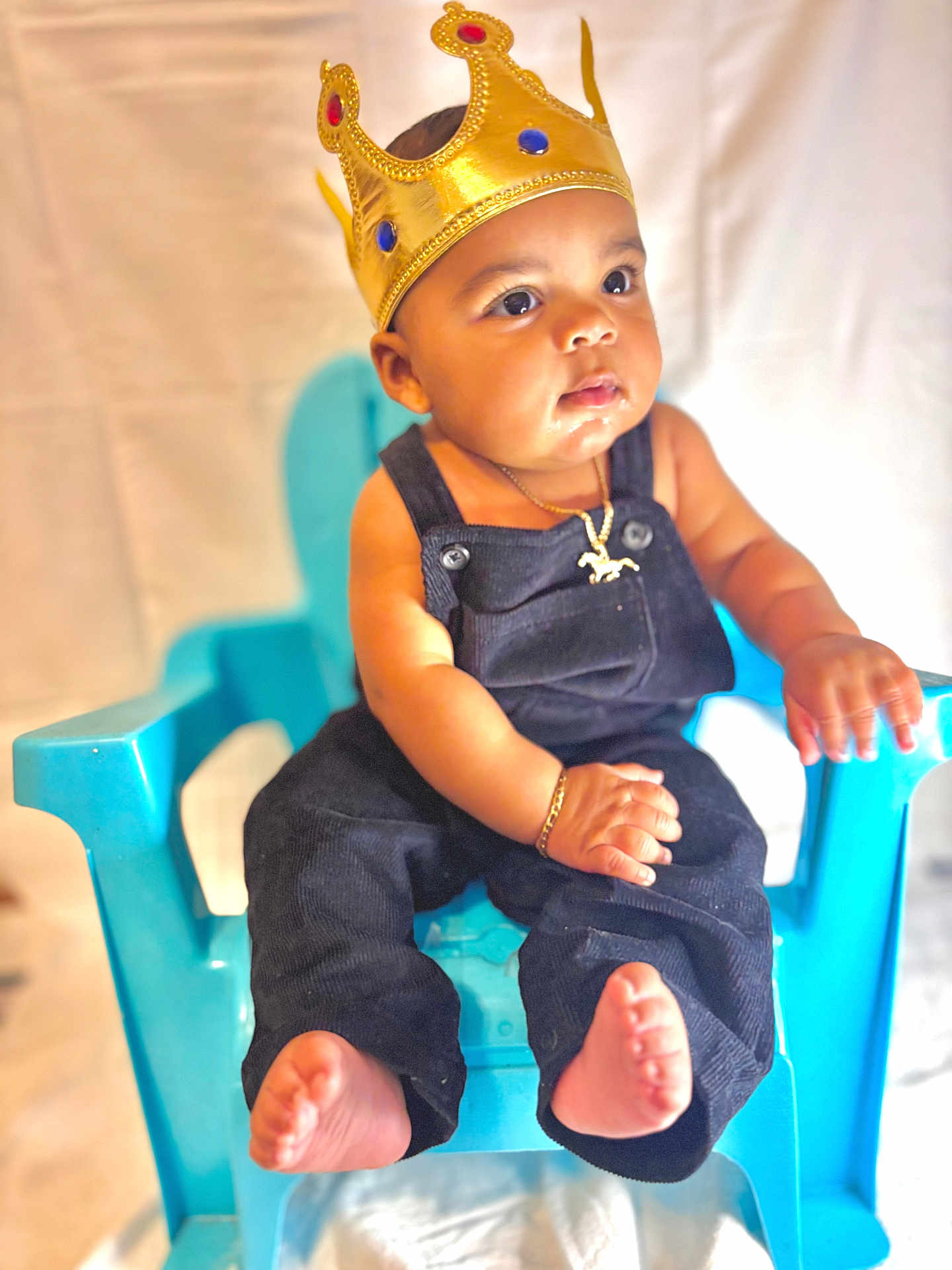 Kylian is registered to the contest to win money with this photo: baby, child, crown, blue_chair, overalls, jewelry, golden_crown, portrait, cute, sitting, person, indoor, infant, baby_clothes, bracelet, necklace, expression, skin, foot, hand