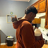 Nova is registered to the contest to win money with this photo: cat, kitten, person, man, hoodie, kitchen, indoors, pet, animal, feline, backpack_cat, curious, sitting, portrait, wooden_cabinets, stove, countertop, trash_can, hardwood_floor, home