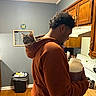 cat, kitten, person, man, hoodie, kitchen, indoors, pet, animal, feline, backpack_cat, curious, sitting, portrait, wooden_cabinets, stove, countertop, trash_can, hardwood_floor, home