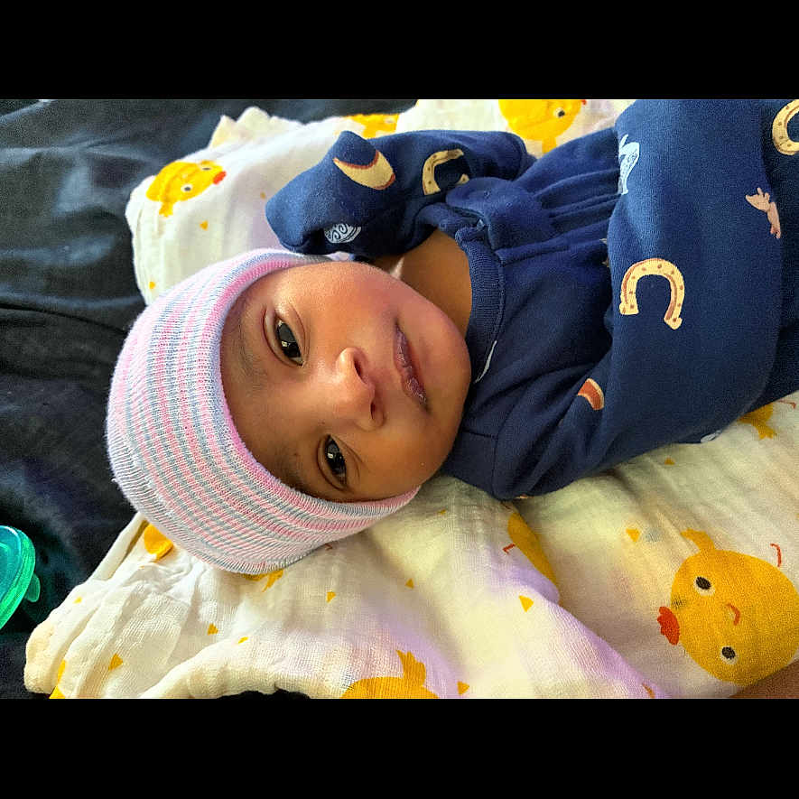 Kmazi Edwards is registered to the contest to win money with this photo: baby, infant, child, portrait, lying_down, blanket, knit_hat, winter_clothing, navy_outfit, striped_hat, duck_pattern, bed, indoor, face, eyes, looking_at_camera, calm, cute, textile, photography