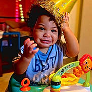 Malikai joined the competition — help win amazing prizes! toddler, party_hat, baby_walker, toy, smile, child, indoor, curly_hair, blue_shirt, colorful, playful, happy, face, person, furniture, background, hand, expression, celebration, home