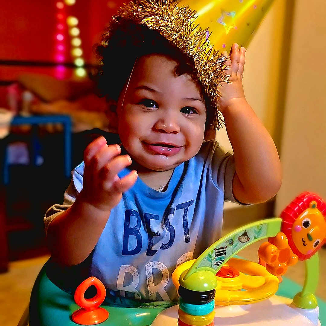 Malikai joined the competition — help win amazing prizes! baby_walker, background, blue_shirt, celebration, child, colorful, curly_hair, expression, face, furniture, hand, happy, home, indoor, party_hat, person, playful, smile, toddler, toy