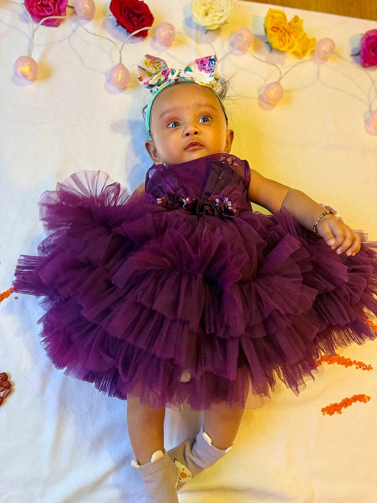 Saanvi is registered to the contest to win money with this photo: baby, child, purple_dress, headband, tutu, lying_down, blanket, flowers, fairy_lights, cute, infant, socks, bracelet, indoor, portrait, adorable, young_child, smiling, decor, celebration