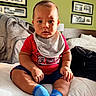 baby, child, infant, portrait, sitting, bed, bedroom, bib, red_shirt, blue_socks, clothing, smiling, face, indoors, luggage, backpack, framed_pictures, furniture, home, photography